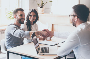 Good deal! Cheerful young man bonding to his wife while shaking hand to man sitting in front of him at the desk